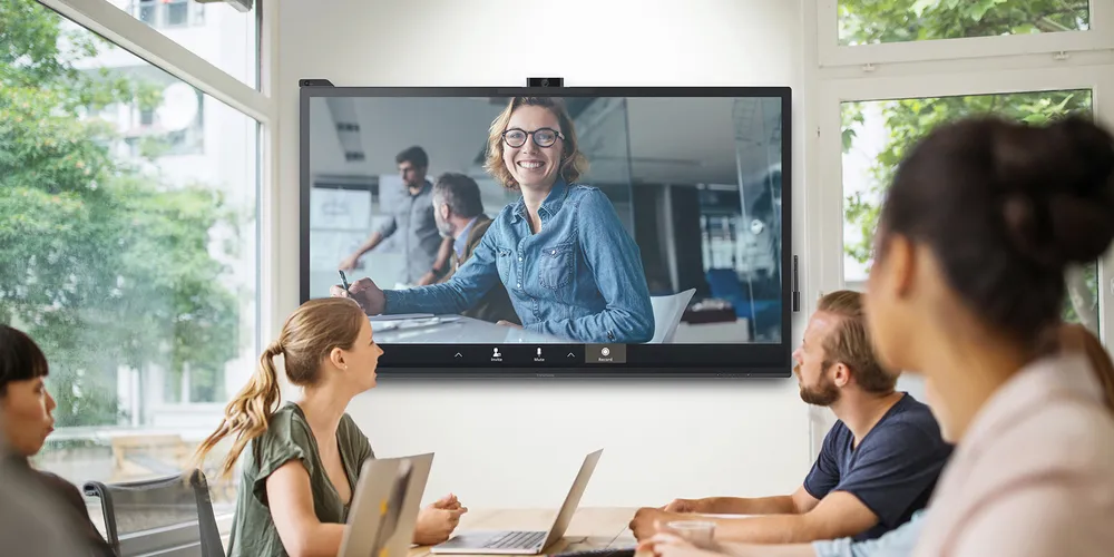 Employees with laptops sitting in a meeting room using a ViewBoard to collaborate with a coworker.