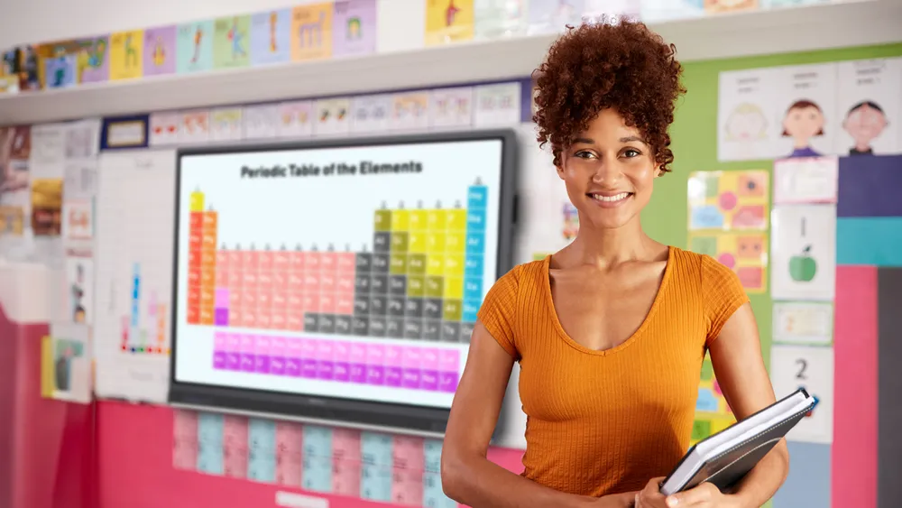 a teacher standing in a classroom.  a viewboard is visible on the wall behind her with the periodic table of elements displayed.