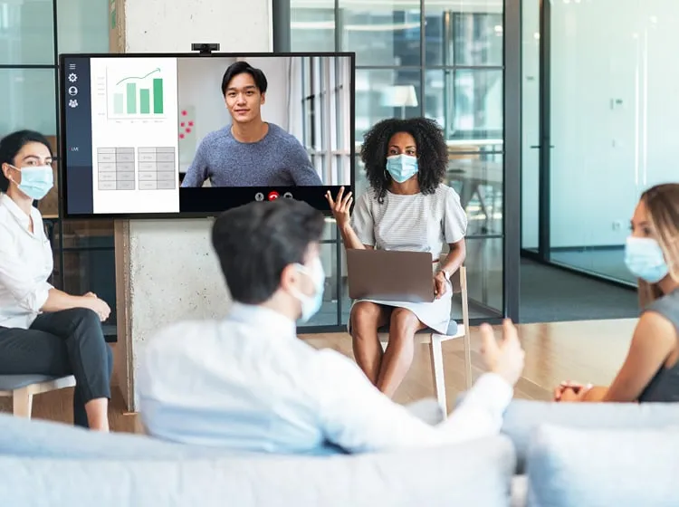 Two people give a presentation in front of their colleagues, while one person joins via video call on the interactive display.